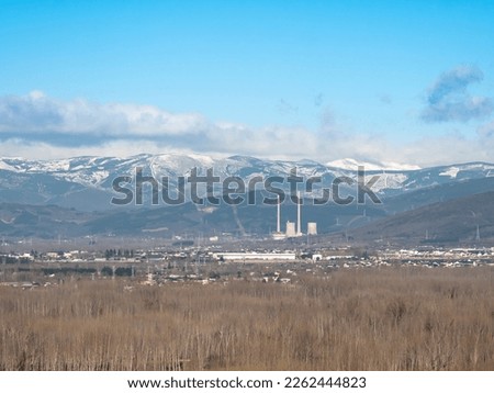 The Compostilla II thermal power plant stands out among the buildings in this view, located between the snow-capped Bierzo mountains with clouds around a sunny winter day and a nice blue sky