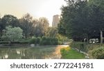 A comprehensive vertical view of a beautiful park landscape featuring a calm pond, diverse tree species, and well-maintained shrubs. People can be seen relaxing in the distance, illustrating the social and ecological value of public green spaces. The warm evening light casts long shadows and highlights the various textures of the flora, making this an ideal shot for tourism marketing, lifestyle blogs, and social media content about park life and recreation.