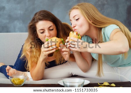 compulsive overeating, mindless snacking, sedentary lifestyle, hunger, pleasure, enjoyment. Two young women sitting on coach greedily engorging burgers and chips
