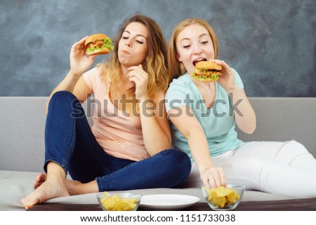 compulsive overeating, mindless snacking, sedentary lifestyle, hunger, pleasure, enjoyment. Two young women sitting on coach greedily devouring burgers and chips
