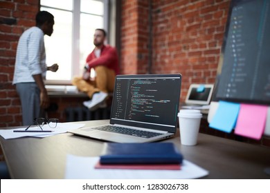 Computer algorithm on laptop display and some other objects on table on background of technicians discussing principles of work