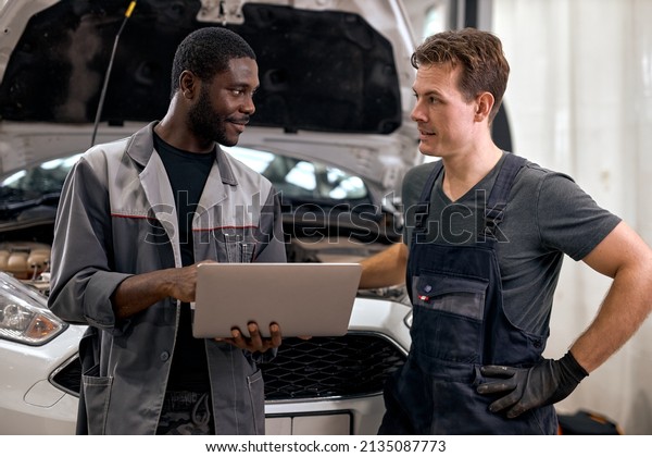 Computer Car Tuning. two Smiling auto mechanic\
testing a car engine connected to laptop. in auto repair shop.\
diverse men in uniform having conversation, discussion.\
cooperation, team work