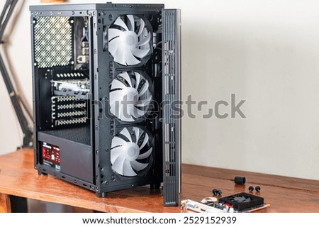 A computer case with three large cooling fans visible, partially open to show internal components. The case is placed on a wooden surface with some additional hardware co