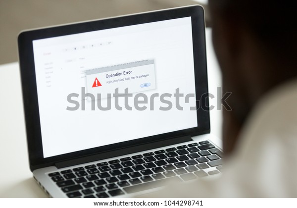 Computer error failure concept, african man\
using laptop with application failure message on screen, bad\
software pc app crash, email malware, data loss and recovery, rear\
view over the shoulder