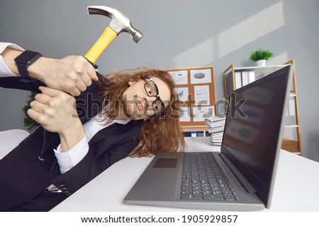 Computer failure, error, stress at work concept. Mad aggressive impulsive crazy adult guy breaks down laptop device screen with hammer. Funny angry nervous office worker smashing monitor and keyboard