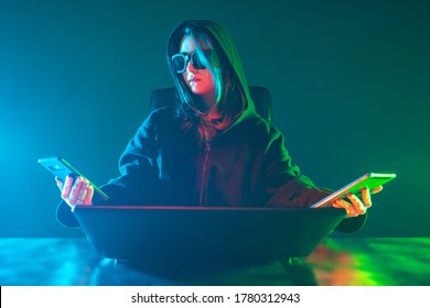 Computer geek woman near laptop. Geek girl holds two phones in her hands. Woman in a neon light. Girl student geek in a dark room. Student in the hood next to a computer. Concept - computer genius.