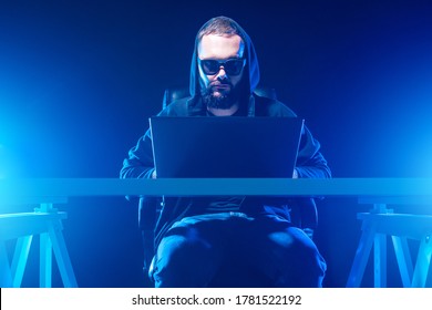 Computer Genius. Computer Geek With A Hood Works Until Late. Computer Geek In A Dark Room. Guy With Laptop Illuminates Blue Light. Geek Man In Sunglasses And A Hood. Man With A Laptop Sits At A Table