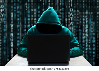 A computer hacker in the hood, with a hidden face, sits in front of a laptop, against a background of binary code. Prosperity of cybercrime, virus outbreaks and online attacks