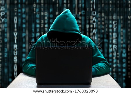 A computer hacker in the hood, with a hidden face, sits in front of a laptop, against a background of binary code. Prosperity of cybercrime, virus outbreaks and online attacks