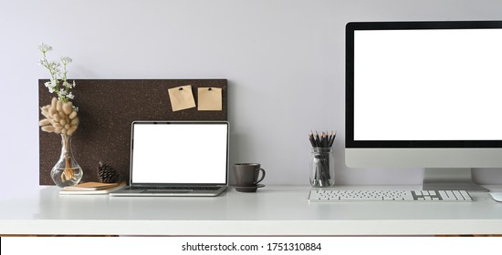 Computer Laptop With Blank Screen Putting On Working Desk That Surrounded By Computer Monitor, Wireless Mouse, Keyboard, Pencil Holder, Coffee Cup, Pine Cone, Notebook, Flowers In Bottle.