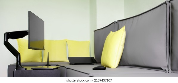 Computer monitor is installed next to gray loft sofa, on small table, using gas-lifted metal swivel bracket. Next to laptop, graphic tablet and mouse. Home workplace for designer or stoker. Daylight