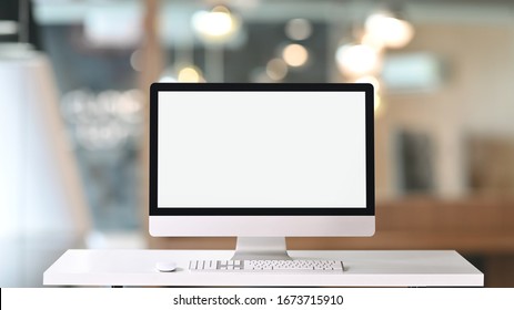 Computer monitor with white blank screen putting on white working desk with wireless mouse and keyboard over blurred vintage office as background.