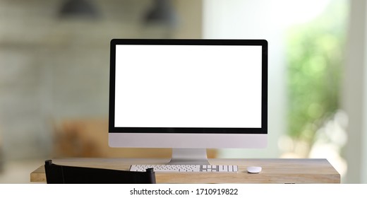 Computer Monitor With White Blank Screen Putting On Workspace Wooden Desk With Keyboard And Mouse Over Blurred Living Room As Background.