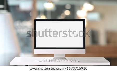 Computer monitor with white blank screen putting on white working desk with wireless mouse and keyboard over blurred vintage office as background.