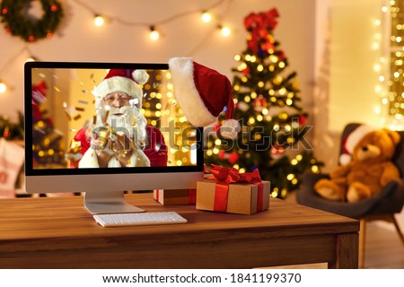 Computer on table in cozy room with hanging red hat and with Santa Claus on screen blowing golden confetti, sending love, wishing Merry Christmas and Happy New Year online and making miracle come true