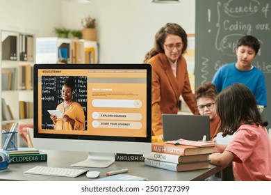 Computer with online learning course on screen at table in school classroom and group of children with female teacher in blurred background copy space