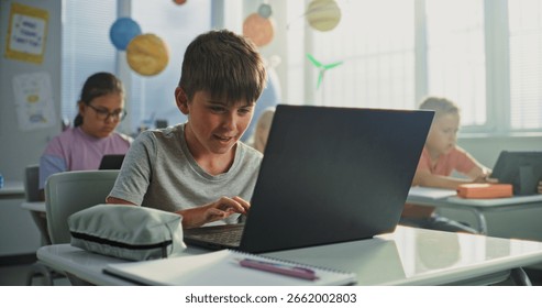 Computer Science Lesson: Primary School Boy Sitting at Desk, Using Laptop in Classroom. Group of Boys and Girls Learning Basic Programming Languages and Internet Safety on Digital Tablets, Laptops.