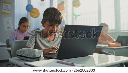 Computer Science Lesson: Primary School Boy Sitting at Desk, Using Laptop in Classroom. Group of Boys and Girls Learning Basic Programming Languages and Internet Safety on Digital Tablets, Laptops.