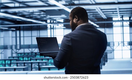 Computer scientist in server farm using laptop to patch network issues leading to slow data transfer rates. IT expert in data center establishment fixing errors affecting supercomputers, camera A