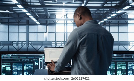 Computer scientist in server room coding on laptop, developing disaster recovery plans. African american man in data center testing failover scenarios using notebook, camera B