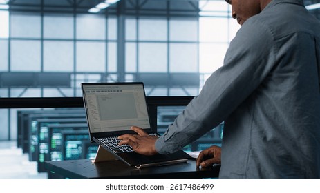 Computer scientist in server room coding on laptop, developing disaster recovery plans. African american man in data center testing failover scenarios using notebook, camera A