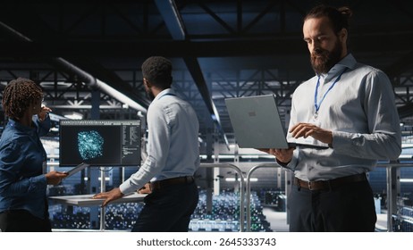 Computer scientists on data center industrial platform analyzing performance outputs on laptop. Server farm IT experts collaborating on finetuning rigs configurations,