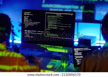 Computer screen with decoded data and two young contemporary programmers sitting by desks and developing new programs