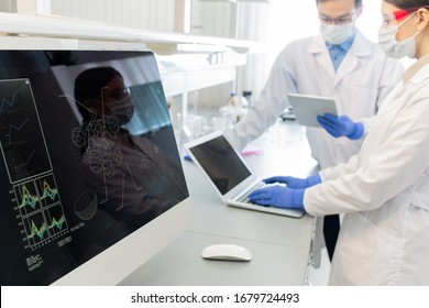 Computer Screen With Molecule Model On Background Of Two Lab Workers In Whitecoats, Masks And Gloves Studying Online Information