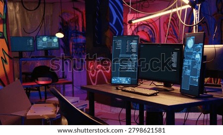 Computer screens showing malware development code in rundown underground hideout with graffiti painted on walls, PC system equipment used for cybercriminal activity in neon lit place.