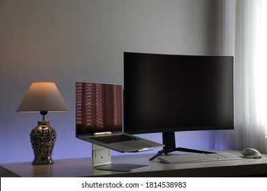 Computer Workstation Ideal For The Home Office Consisting Of A Gray Laptop On A Stand, A Large Curved And Black Monitor, A White Lamp, A White Keyboard And A White Optical Mouse With Backlighting