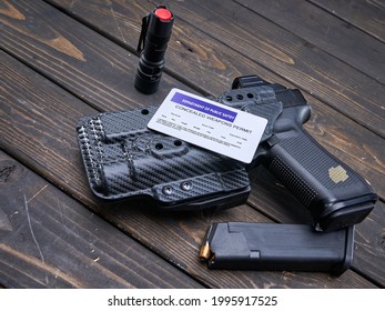 Concealed Carry Permit Sitting On Top Of A Holstered Handgun With Spare Magazine And Flashlight On Wood Table. 