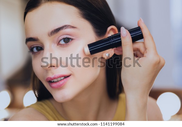 Concealer stick. Charming beautiful woman using\
concealer stick while putting makeup\
on
