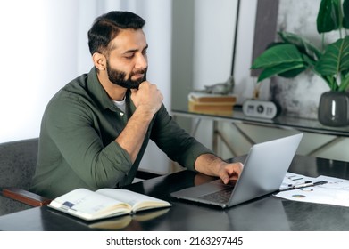 Conceived serious bearded indian or arabian guy in casual clothes, office worker or freelancer, sits at the desktop in the modern office, looks into the laptop intently, thinks about the project - Shutterstock ID 2163297445