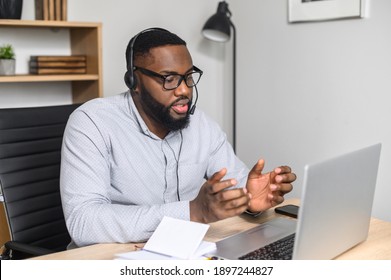 Concentered Young African-American Student Presenting His Project Online On The Laptop, Wearing Glasses And Headset, Making Notes, Focused Speaker Briefing The Sales Team On A Video Call