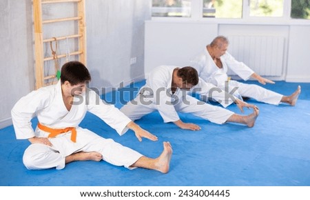 Concentrated adolescent jiu-jitsu fighter doing stretching exercises on mat, warming up body muscles before training during group martial arts class