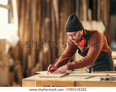Concentrated adult woodworker marking wooden piece with pencil while working at workbench in joinery studio