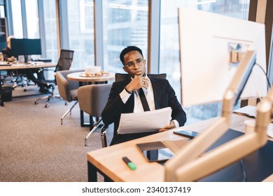 Concentrated African American male entrepreneur in formal suit holding statistic papers and analyzing information on computer screen at wooden desk in modern light office - Powered by Shutterstock