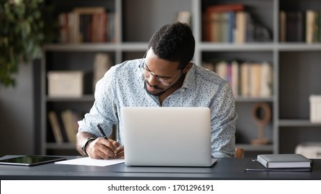 Concentrated African American male worker sit at desk handwrite watching webinar or training on laptop, focused biracial man make notes busy studying working on computer at workplace in office