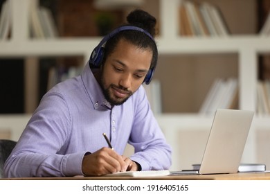 Concentrated African American man in earphones write make notes study online on computer from home. Mixed race ethnicity male student in headphones handwrite watching webinar on laptop.
