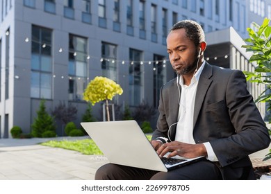 Concentrated african american man in suit sitting near office center outside with laptop and headphones, and having online interview, talking on video call.