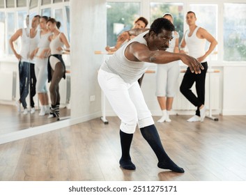 Concentrated African American middle-aged male ballet dancer performing solo routine during group practice in light spacious choreography studio, executing graceful artistic movements.. - Powered by Shutterstock