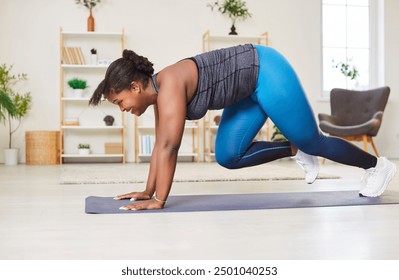 Concentrated African American young woman doing intense workout in living room at home, performing mountain climber exercise in plank pose on exercise mat. Smiling woman training cardio indoors. - Powered by Shutterstock