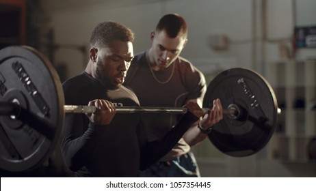 Concentrated African-American sportsman training bicep with heavy barbell under control of professional coach in gym.