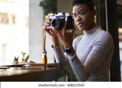 Concentrated Afro American Female Photographer Holding Vintage Camera Taking Picture Of Modern Designed Interior Of Coffee Shop Working Sitting Near Window At Wooden Table With Notebook And Stationery