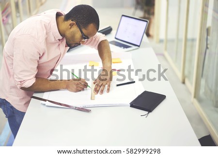 Concentrated afro american skilled architect creating graphic of construction drawing detailed outlay by hand before using specialist computer-aided design applications on laptop with mock up screen