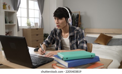 Concentrated Asian Chinese Male Student Learning From Home With Headphones Is Practice Speaking And Writing Notes While Taking Online Language Class On Laptop In Bedroom