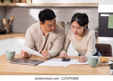 Concentrated Asian Couple Sitting At Table Full Of Papers, Laptop, Coffee Mugs, Counting Monthly Expenses, Using Calculator, Short Of Money, Kitchen Interior. Economy, Family Budget Concept