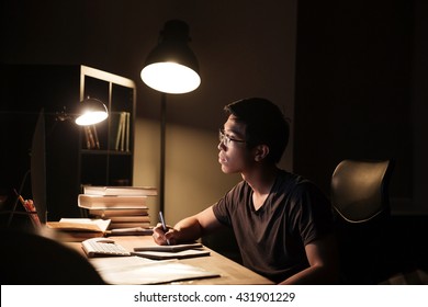 Concentrated Asian Young Man In Glasses Using Computer For Studying And Writing In Notepad In The Evening At Home