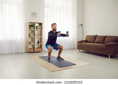 Concentrated Athletic Man Trains Squatting With Dumbbells During Workout At Home. Beginner Or Amateur Holds Small Dumbbells In Outstretched Hands And Squats In Apartment. Home Sport Concept.