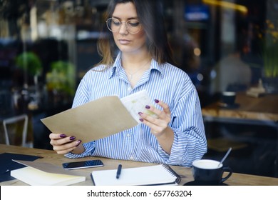 Concentrated attractive business woman in trendy eyewear opening envelope with copy space for advertising checking financial papers, administrative manager receiving letter on mail sitting in cafe